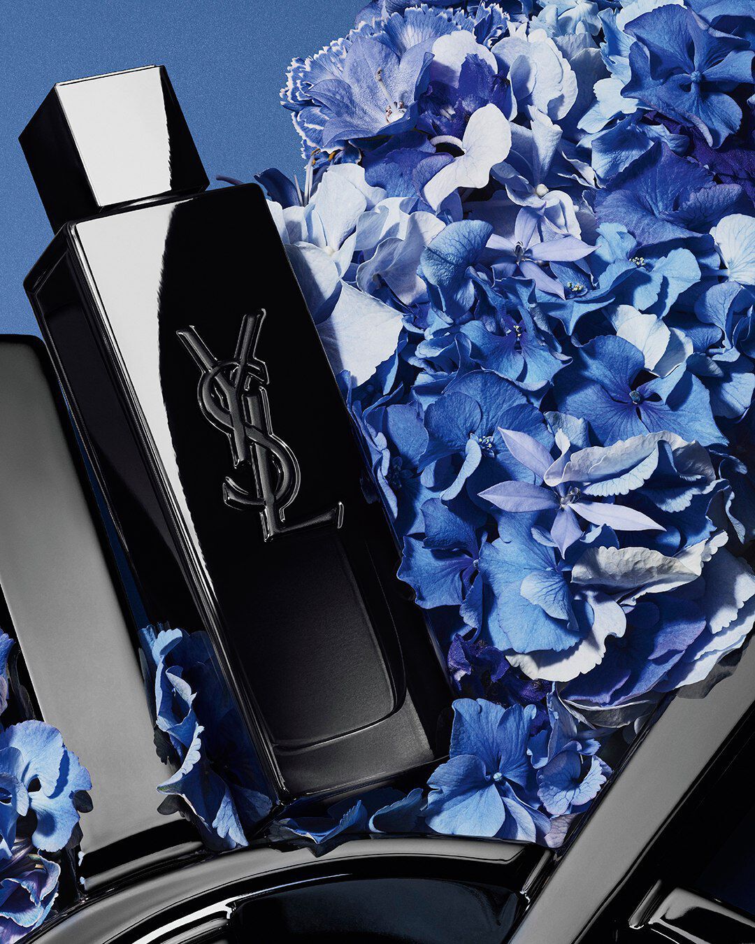 YSL MYSLF Eau de Parfum bottle on a glossy black YSL logo, with vibrant blue hydrangeas, against a deep blue background.