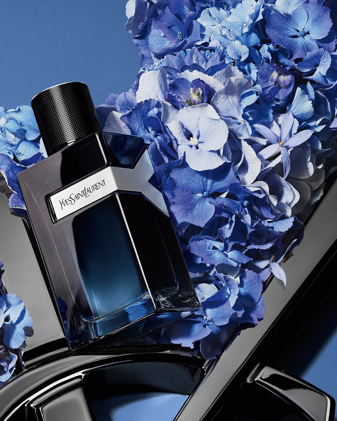 YSL Y Eau de Parfum bottle on a glossy black YSL logo, surrounded by vibrant blue hydrangeas, against a deep blue background.