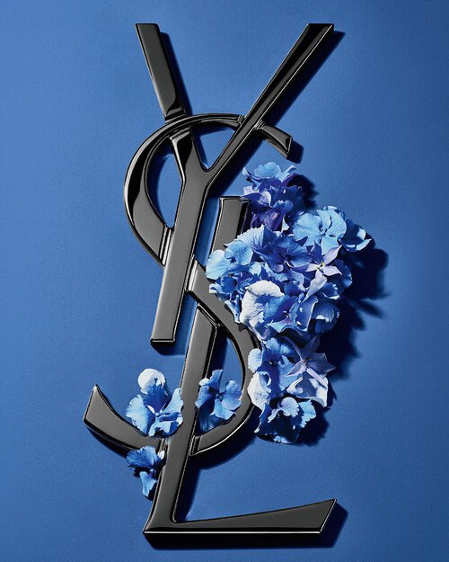 Black YSL Cassandre logo adorned with vibrant blue spring flowers, set against a serene blue background.